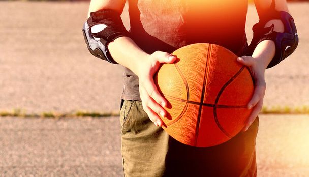 Πορτοκαλί μπάλα μπάσκετ στα χέρια ενός εφήβου στο φόντο του γηπέδου μια καλοκαιρινή μέρα. Κοντινό πλάνο. Αντιγραφή χώρου στα αριστερά - Φωτογραφία, εικόνα