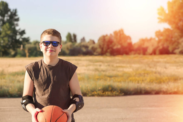 Smiling boy in blue T-shirt and sun glasses plays basketball on the city playground. Active teenager enjoying outdoor games with an orange ball. Hobby, active lifestyle, sports. Copy space on the righ - Zdjęcie, obraz