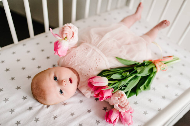 Greeting for the mother with the newborn baby girl who holding the flower and lying on a bed with a bouquet of pink tulips. Women's Day. Mother's Day. - Photo, image