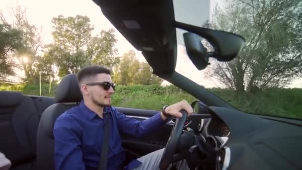 stylish young man in black sunglasses rides in a cabriolet. Caucasian brunette man with a beard drives a convertible. Car with an open roof on a background of nature and trees. Happy sings. Sunset - Záběry, video