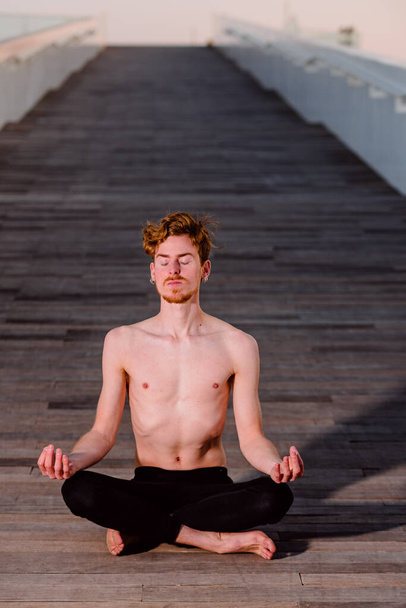 Joven practicante de yoga entrenando sus poses, piernas cruzadas sentada en un suelo de madera en el parque de la ciudad al atardecer, pose de loto
. - Foto, Imagen