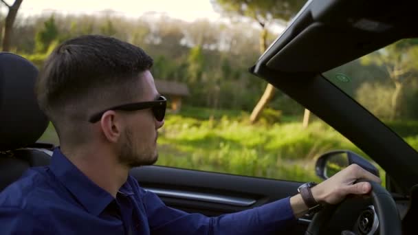 stylish young man in black sunglasses rides in a cabriolet. Caucasian brunette man with a beard drives a convertible. Car with an open roof on a background of nature and trees. Happy sings. Sunset - Filmati, video