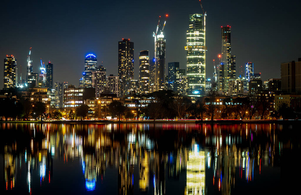 dłuższą ekspozycję fotografii z odbiciami w nieruchomej wodzie. Odbicia panoramy miasta Melbourne o zmierzchu w spokojnej wodzie jeziora Albert Park - Zdjęcie, obraz