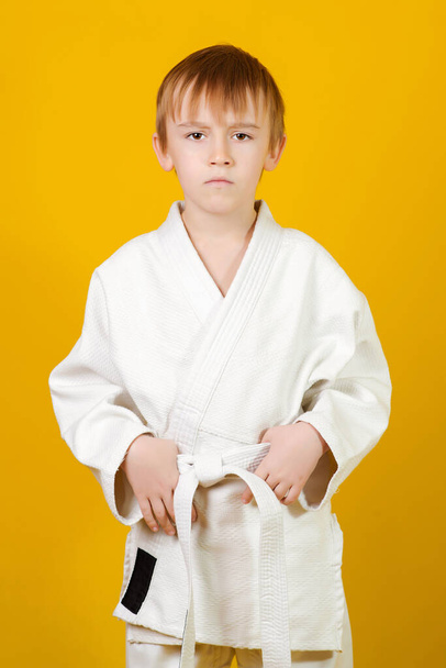 Cute serious boy in white kimono over yellow background. Sports since childhood, discipline, first place, victory. Little boy with white belt on kimono. Kids sport, martial arts, karate. - Photo, Image