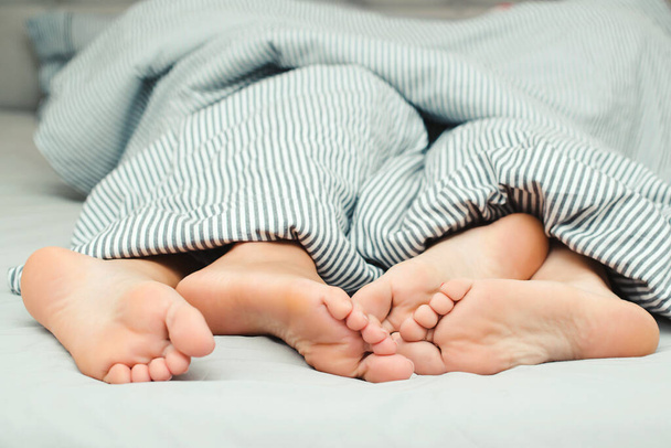 Feet of a young couple lying on the bed at honeymoon. Passionate love. Love lying in bed in hotel. Close up legs under stripes blanket. Sensual and intimate moment of lovers. Morning time together. - Photo, Image