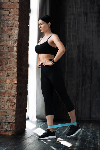 Full length portrait of a young woman in black sportswear who trains calf muscles with an elastic expander to pump up leg muscles on the floor in a loft - Foto, afbeelding