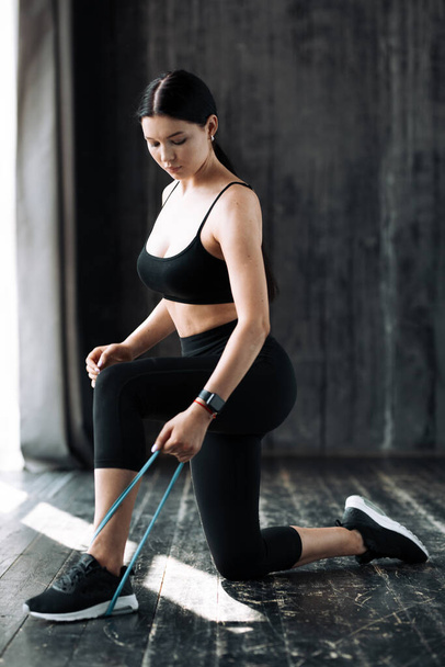 Full-length A young woman is kneeling and pressing down an elastic expander with her leg to pump up the muscles of her arms on the floor in a loft - Фото, изображение