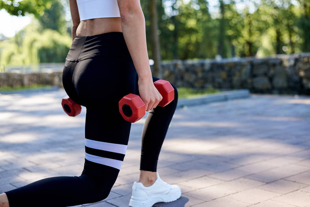 Young blond woman, wearing white top and black leggings,doing power exercises in park in summer.Training process outdoors. Close-up picture of female legs, doing lunges, holding red fitness weights. - Φωτογραφία, εικόνα