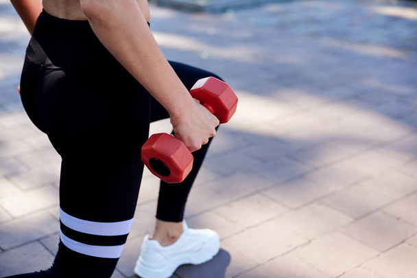 Young blond woman, wearing white top and black leggings,doing power exercises in park in summer.Training process outdoors. Close-up picture of female legs, doing lunges, holding red fitness weights. - 写真・画像