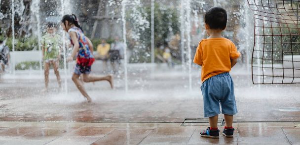 Uroczy chłopiec azjatycki (1-latek) stojący i cieszący się widokiem publicznych fontann na świeżym powietrzu po raz pierwszy tryska wodą. Tajlandia bawi się i przestrzeń zabawy dla dzieci. Koncepcja wakacji i wakacji. - Zdjęcie, obraz