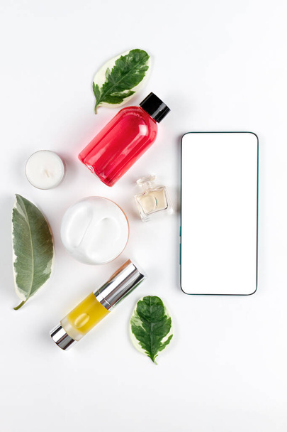 Cosmetics. On a white background are shower gel, oil in a tube, candle, cream and perfume, next to the phone. Flat lay. Mock up. Concept of modern technology and beauty. Vertical. - Фото, зображення