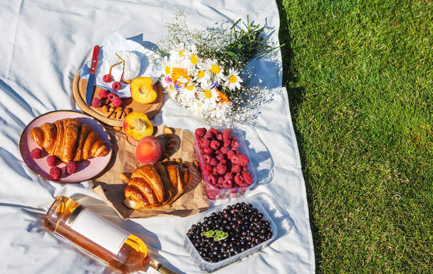 Letni piknik ekologiczny na trawie z rogalikiem, brzoskwiniami, jagodami i winem na białym obrusie. Widok z góry. - Zdjęcie, obraz