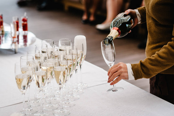 Man hands pouring champagne from a bottle in glass that stand on buffet table. Close up. The man fills the glasses elite drinks standing in three rows. - Fotografie, Obrázek