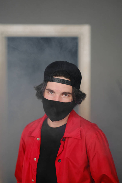 Portrait of a young man of 25-30 years old in a black protective mask, cap and red jacket.  - 写真・画像