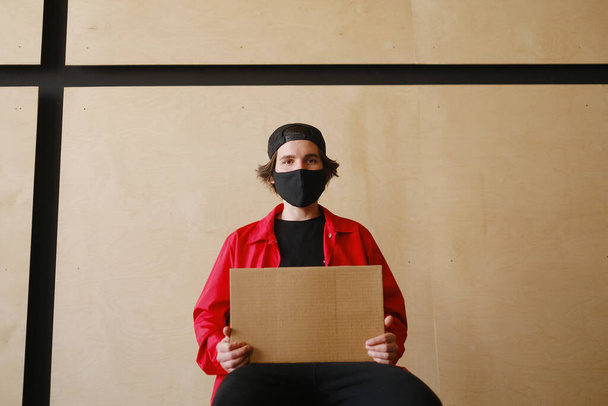 Молодий чоловік 25-30 років в чорній захисній масці, жовтих окулярах, шапці і червоній куртці тримає в руках порожній картонний знак
. - Фото, зображення