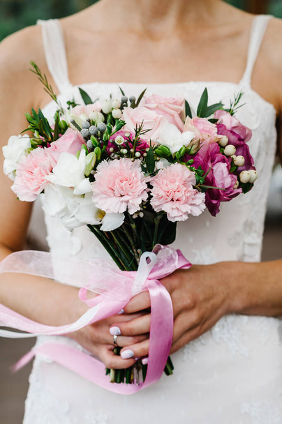 Young beautiful bride in an elegant dress is standing and holding bouquet of pink and purple flowers and greens with ribbon at the wedding ceremony. - Foto, immagini