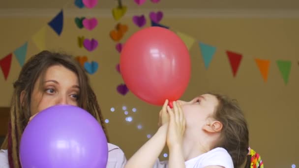 Çocukların doğum günü partisi. Küçük tatlı kız doğum gününü genç annesiyle kutluyor. Rasta desenli balonları şişiriyor. Evinde kağıt çelenk takıyor. - Video, Çekim