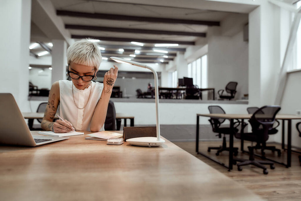 Working on new project. Young, beautiful tattooed business lady in eyewear making some notes while sitting alone in the modern office - Фото, зображення
