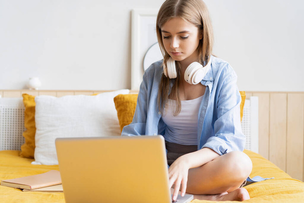 Pretty young student working at home on her laptop computer sitting on her bed. - 写真・画像