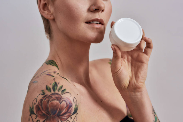 Cropped portrait of beautiful tattooed woman with pierced nose and short hair holding white plastic jar of cream or body lotion isolated over grey background - Фото, изображение