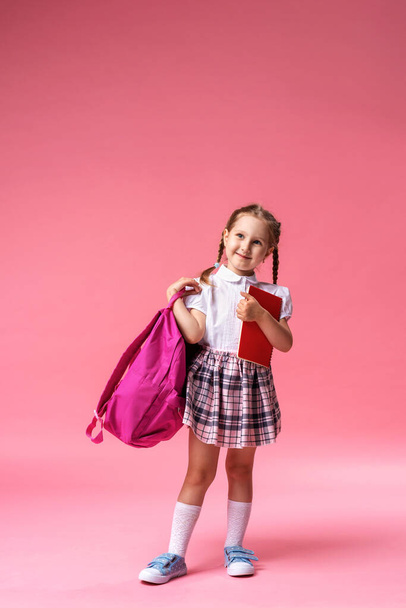 happy smiling little girl in uniform with a backpack and a notebook in her hands poses on a pink background in the Studio. The child goes to school for the first time. Back to school. copy space - Фото, изображение