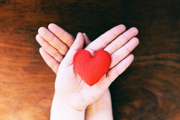 Hart in de hand voor filantropie concept - vrouw houdt rood hart op handen voor Valentijnsdag of doneren hulp geven liefde warmte zorg met houten achtergrond - Foto, afbeelding