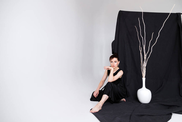 Een vrouw in een zwarte minimalistische jurk op een zwarte stof achtergrond zit in de studio. 35-40 jaar oud - Foto, afbeelding