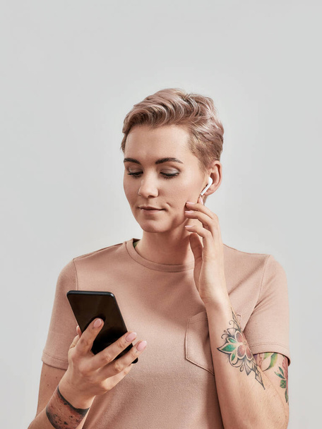 Portrait of tattooed woman with pierced nose and short hair in wireless earbuds or earphones using smartphone isolated over light background - Фото, изображение
