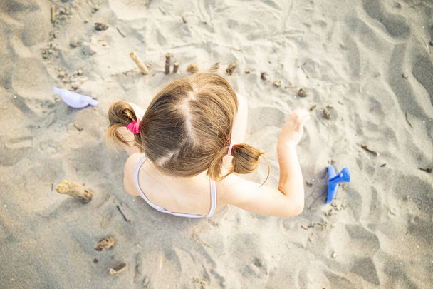 Caucasian child girl playing with sand and driftwoods while sitting on the beach, top view. - Photo, Image