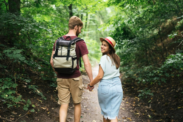 男と女が一緒に木の歩道に沿って、手を握って歩く。バックビュー。幸せなカップル手を握り、森林のパスを歩く。夏はバックパック付きの森の中を歩くハイカー. - 写真・画像
