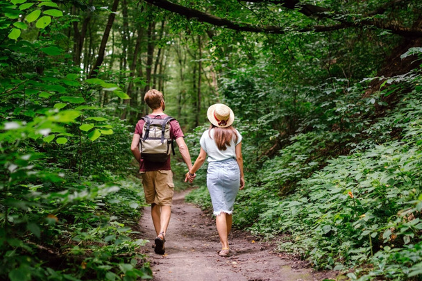 Couple holding hands walking in forest, back view. Adventure, travel, tourism, hike, people. Rear view of two people carrying backpack while walking through wood. Man holds girlfriend by hand on hike. - Photo, image