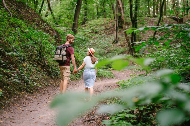 Happy caucasian couple are walking in a dense forest along the path holding hands, rear view. Hikers with backpack looking for place for picnic in wooded area in the summer. Walk outdoors together. - Photo, image