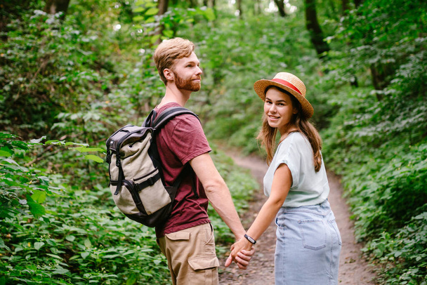 Happy caucasian couple are walking in a dense forest along the path holding hands, rear view. Hikers with backpack looking for place for picnic in wooded area in the summer. Walk outdoors together. - Photo, Image