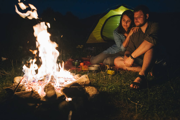 glimlachend vrolijk paar zitten voor het kampvuur koken diner. kampeerconcept. kopieerruimte - Foto, afbeelding