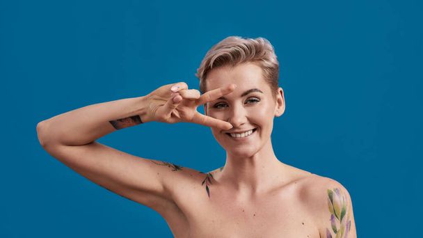 Portrait of a young attractive half naked tattooed woman with perfect skin looking happy, holding hand on eyes and showing victory or peace sign isolated over blue background - Zdjęcie, obraz