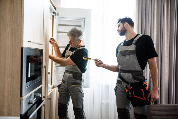 The technical people. Aged repairman in uniform fixing refrigerator in the kitchen, while his colleague helping him, bringing screwdriver. Repair service concept - Foto, afbeelding