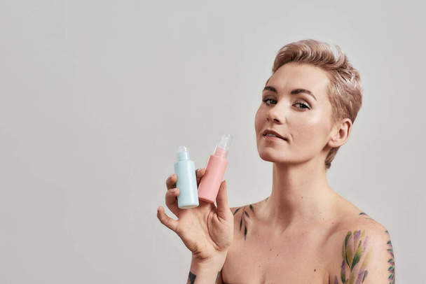 New Beauty. Portrait of beautiful tattooed woman with pierced nose and short hair holding plastic bottles with cosmetic, hair and skin care products, isolated over grey background - Foto, Imagem
