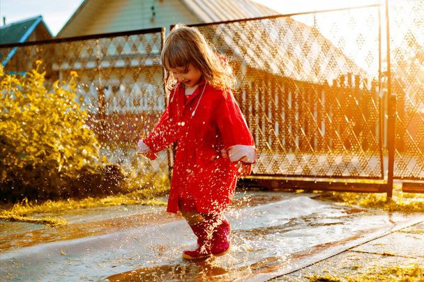 Cute girl in a red jacket is jumping in the puddle.The setting warm summer or autumn sun. summer in the village - Fotó, kép