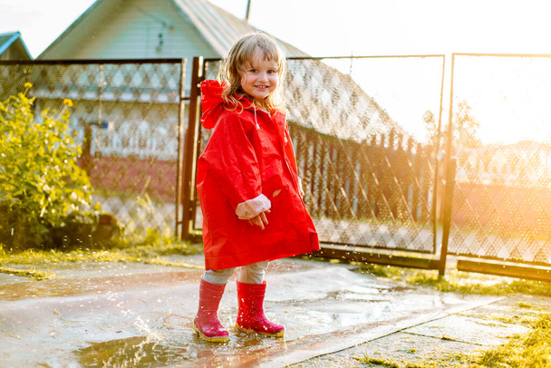 Cute girl in a red jacket is jumping in the puddle.The setting warm summer or autumn sun. summer in the village - Фото, изображение