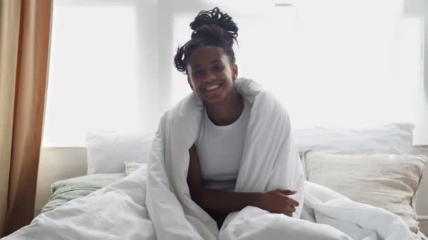 Giovane donna africana felice si siede om letto, guarda la fotocamera e sorride - Filmati, video