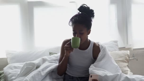 Κοντινό πλάνο του Νεαρή Αφροαμερικανή γυναίκα κάθεται στο κρεβάτι, πίνει πρωινό καφέ και χρησιμοποιεί το τηλέφωνο - Πλάνα, βίντεο
