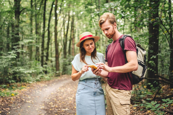 Turyści korzystający z mobilnych GPS-ów w celu uzyskania wskazówek. Szczęśliwa para sprawdza smartfona w lesie podczas podróży z plecakiem. Młoda radosna para korzystająca z mapy GPS, aplikacji geolokalizacji nawigatora. koncepcja miłości i podróży. - Zdjęcie, obraz