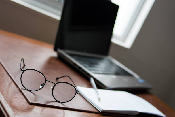 Klasyczne czarne okulary z laptopem i notatnikiem na drewnianym stole z oknem jako tłem   - Zdjęcie, obraz