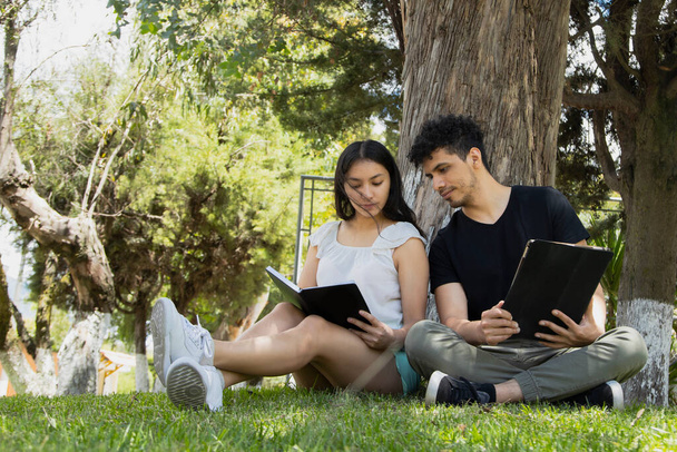 Młoda Latynoska para siedząca pod drzewem i czytająca w parku w słoneczny dzień - młodzi ludzie uczący się przy pomocy tabletu elektronicznego i książki w środku natury - szeroki kadr - Zdjęcie, obraz