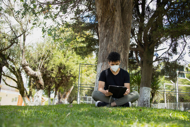 Maskeli İspanyol genç adam parkta oturuyor. Genç adam, doğal bir parkta elektronik tabletle ders çalışıyor. Sosyal uzaklık. - Fotoğraf, Görsel