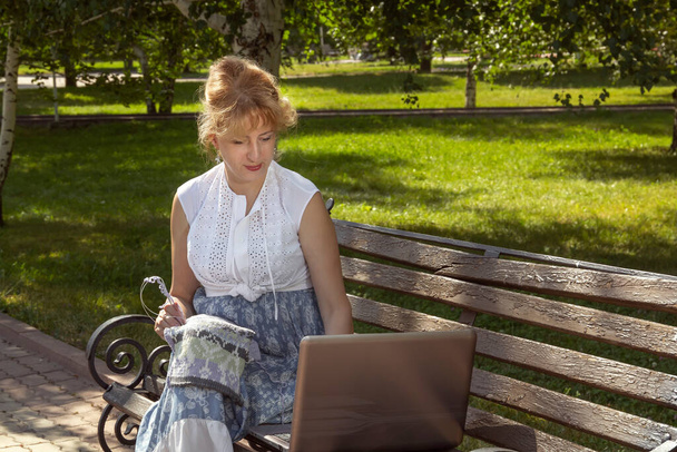 Woman in a park sits on a bench knitting wool clothes on knitting needles and watching lessons or teaching knitting through a laptop. Distance learning. Copy space - Photo, Image