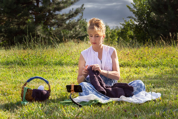 γυναίκα κάθεται στο γρασίδι σε ένα πάρκο πλέξιμο μάλλινα ρούχα για το πλέξιμο βελόνες και ρολόγια μαθήματα ή διδάσκει πλέξιμο μέσω ενός κινητού τηλεφώνου. Εκμάθηση εξ αποστάσεως - Φωτογραφία, εικόνα