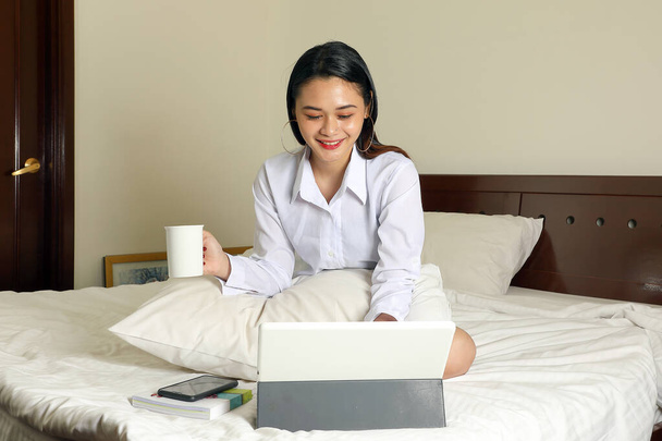Fiatal gyönyörű modern maláj nő ül az ágyban munka tanulmány olvasni írni a hálószobában laptop fül mosoly nézni kamera - Fotó, kép