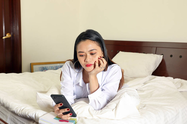 Young beautiful modern Malay woman sit on bed work study read write in bedroom laptop tab book smile look at camera - Photo, Image