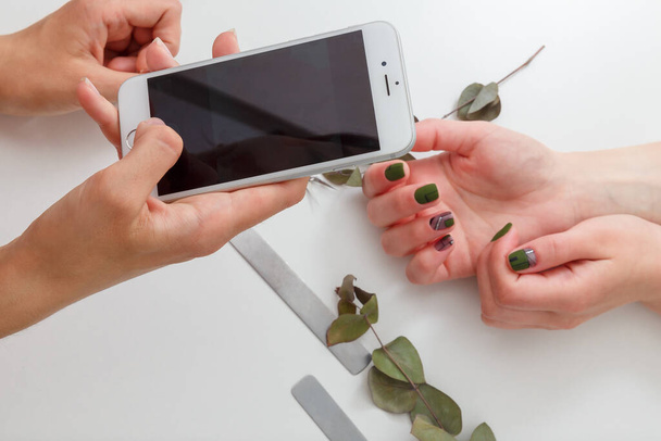 Nagelkünstlerin, die mit dem Smartphone Fotos von ihrer Arbeit macht, Kunden, die ihre Hände gepflegt haben. Grüner Nagellack, Nagelfeilen und Eukalyptuszweige auf weißem Tisch. Frühjahrsmaniküre. - Foto, Bild